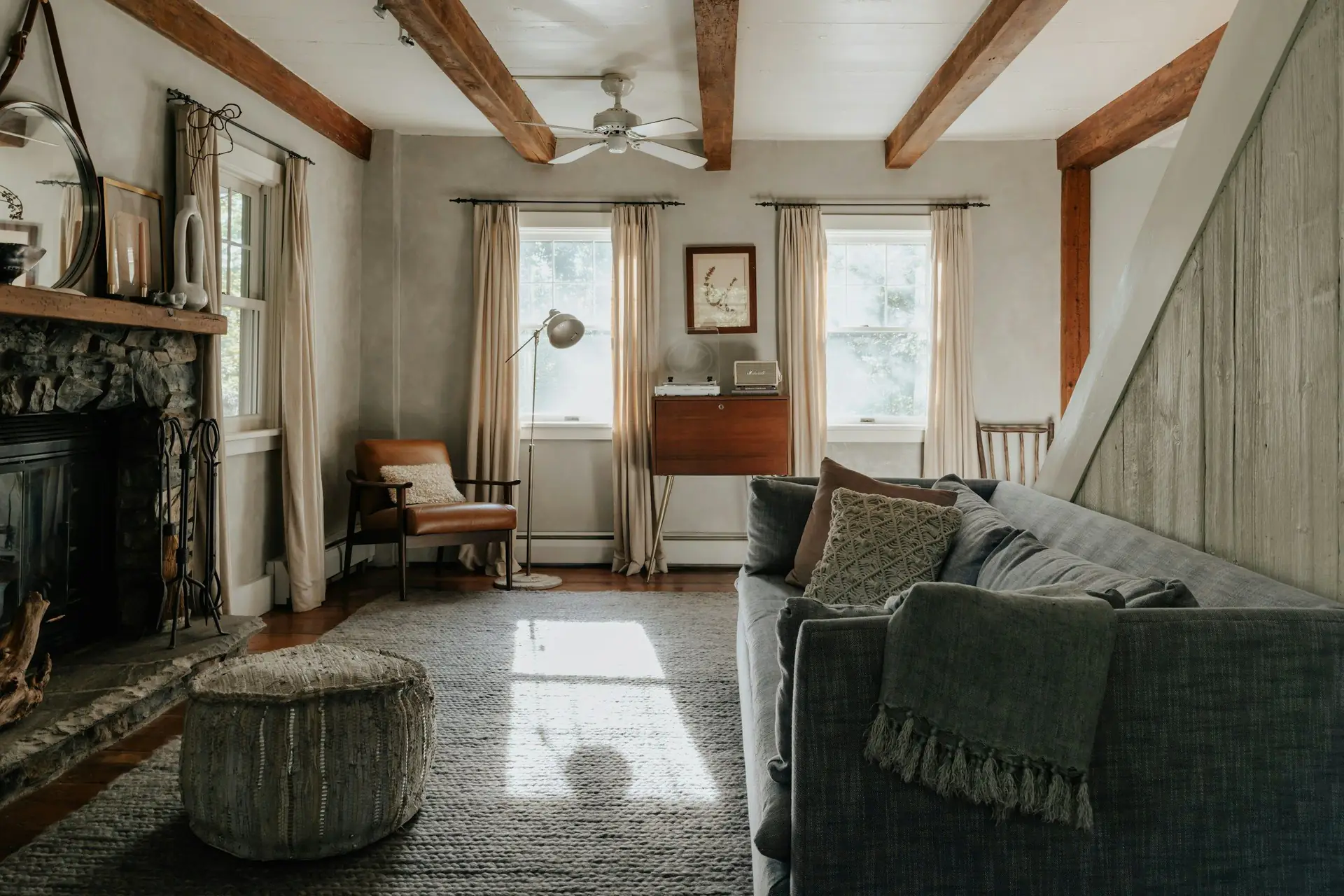 A calm living room with timber beams, a stone fireplace and linen curtains