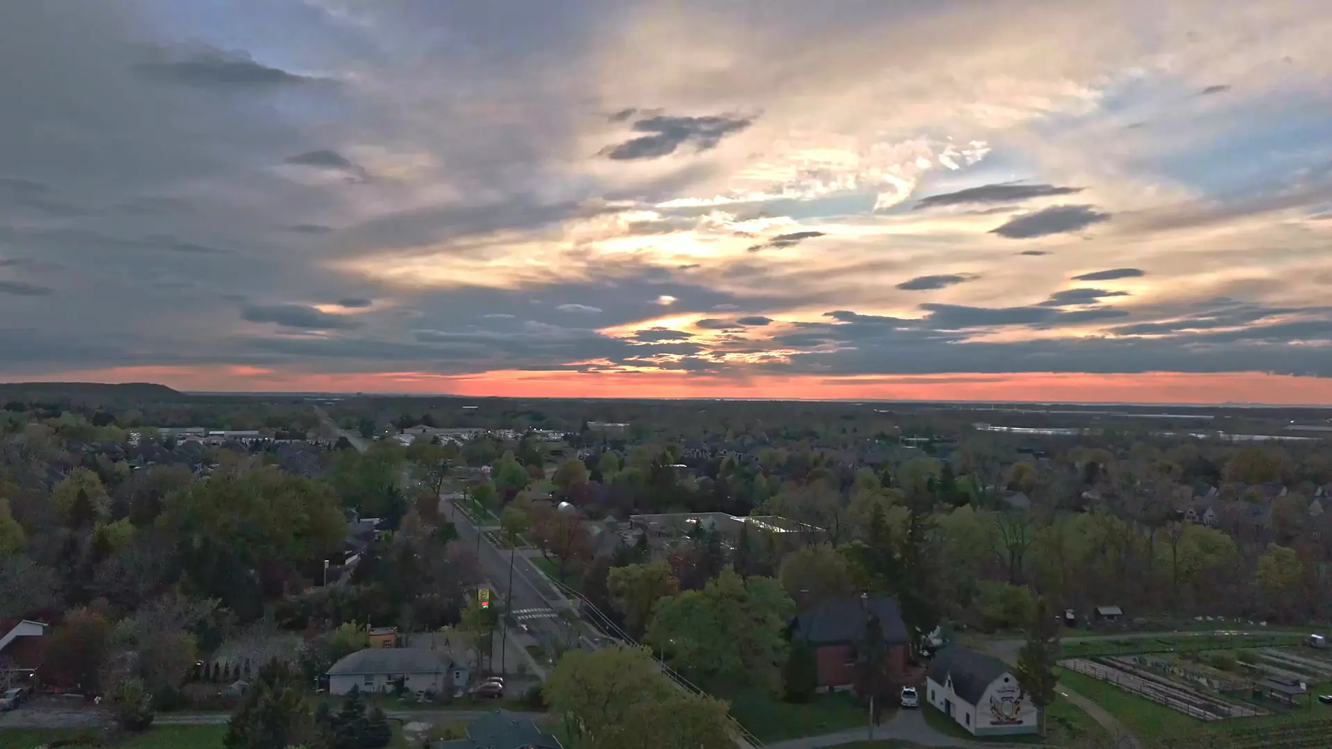 Drone view rising over a leafy neighbourhood of houses and tree-lined streets under a wide pink and gold sunset sky